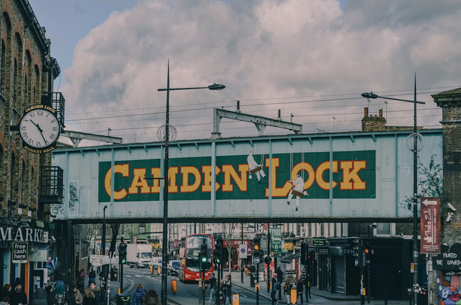 A bustling outdoor scene at Camden Market in London during daytime, featuring multiple green and white umbrellas with the 'Camden Market' logo providing shade to visitors. The market has a brick exter