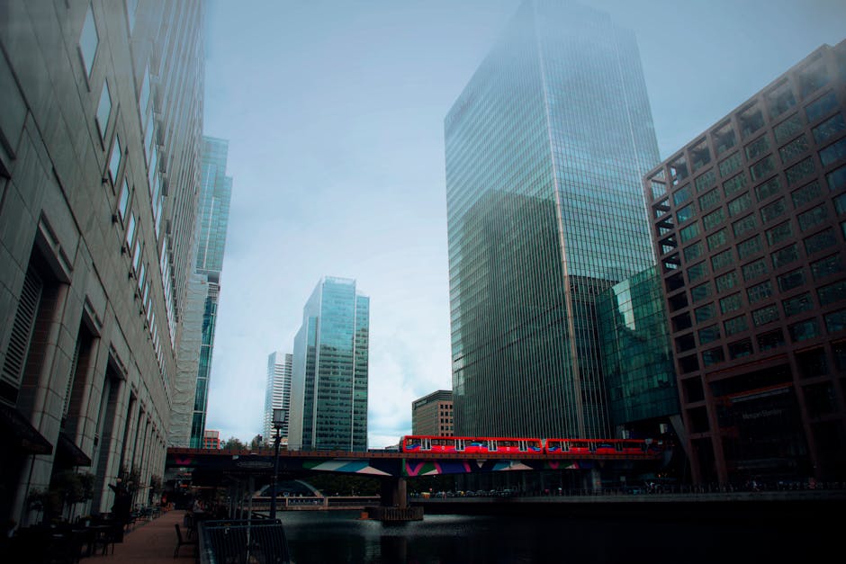 A modern office building with a curved glass façade reflects the surrounding skyscrapers and overcast sky, highlighting the sleek, clean surfaces of the extensive glass windows and steel framework. T